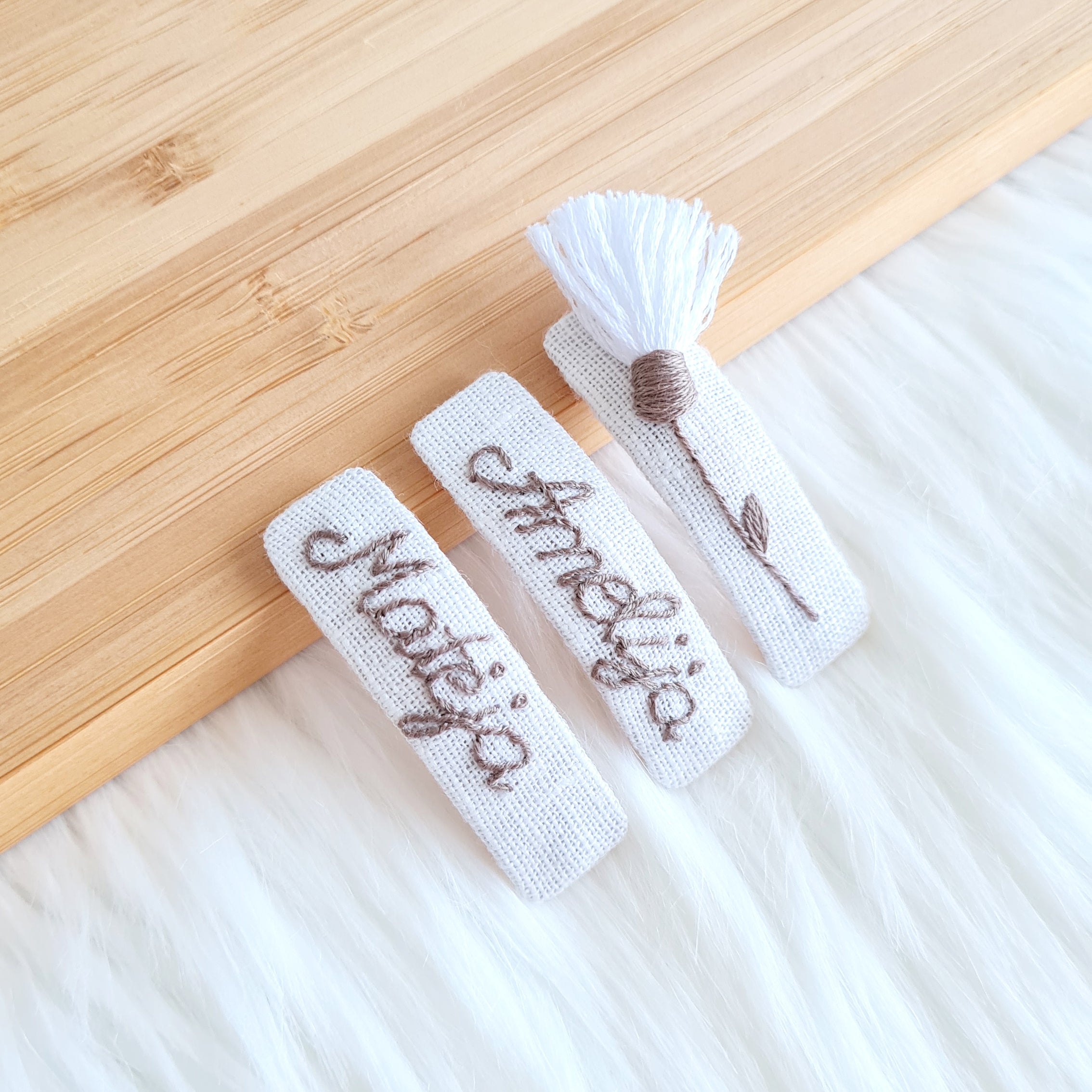 Rankomis siuvinėtų lininių plaukų segtukų komplektas, sudarytas iš trijų balto lino segtukų. Du segtukai su siuvinėtais vardais, o trečias – su siuvinėta smilga ir baltu kutu viršuje.